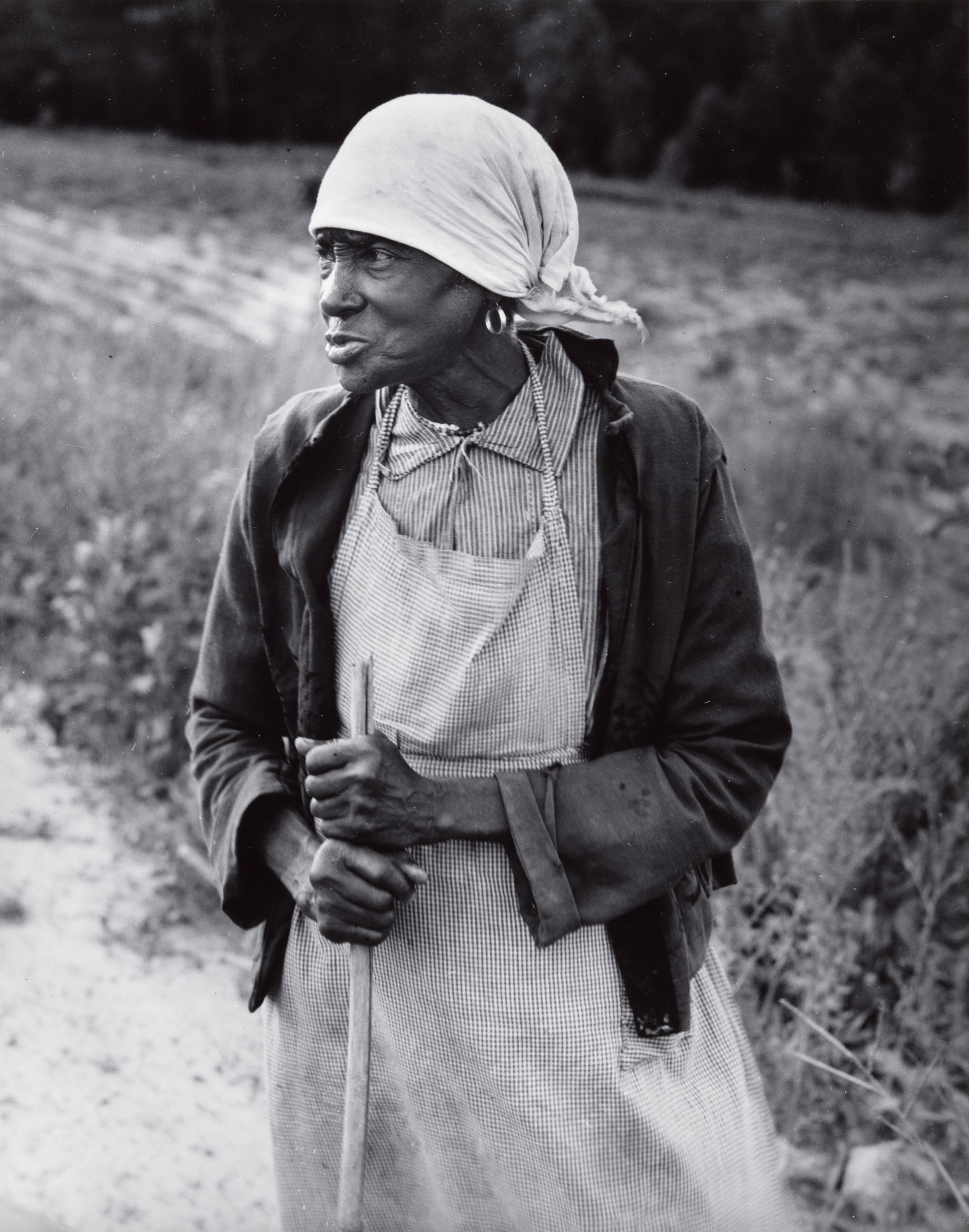 Black-and-white photograph of a Black woman wearing a light-colored headscarf, standing in a field, supporting herself on a wooden staff.