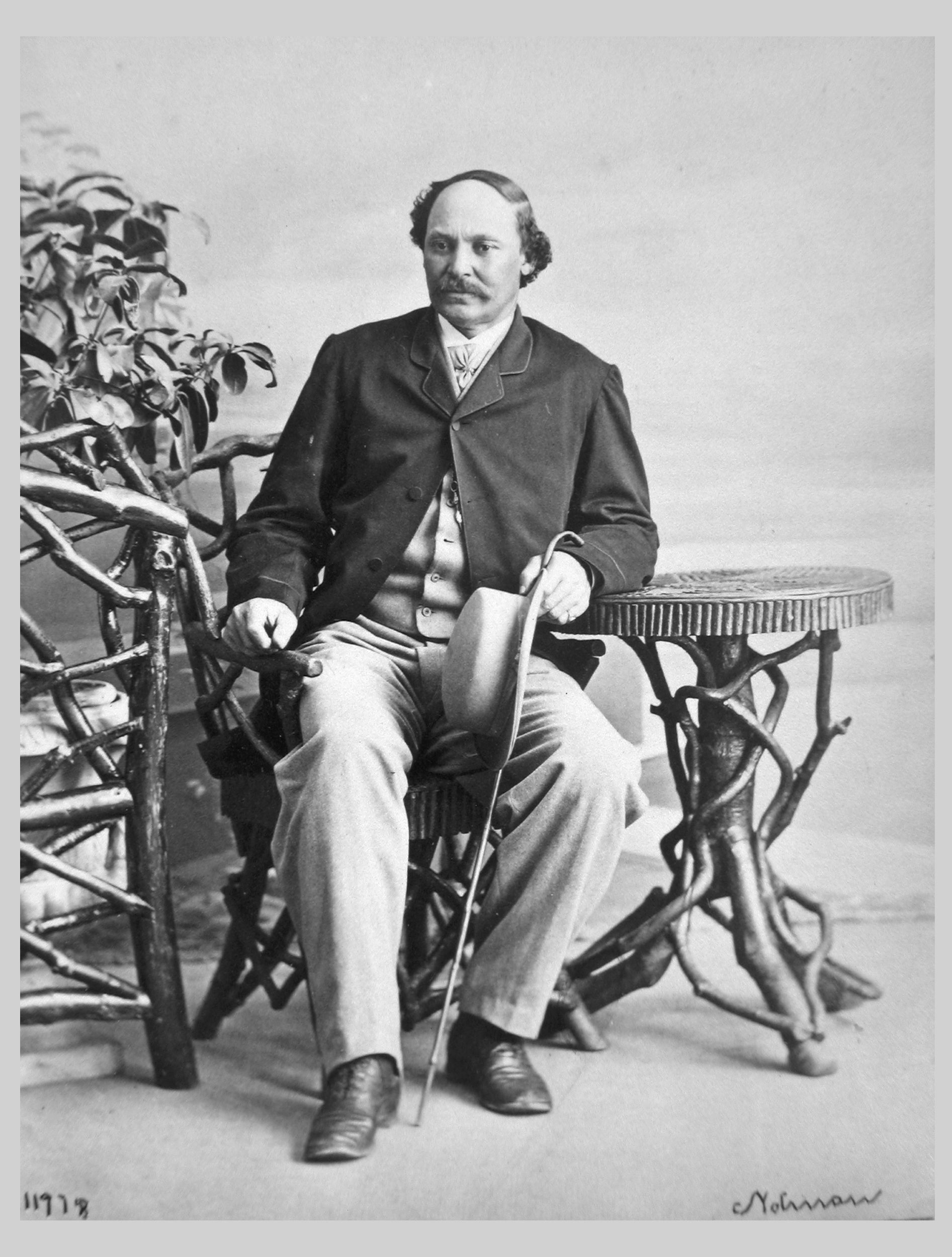 Black-and-white photograph of a white-appearing man seated at a chair and small table made of tree branches, holding a can and straw boater hat.