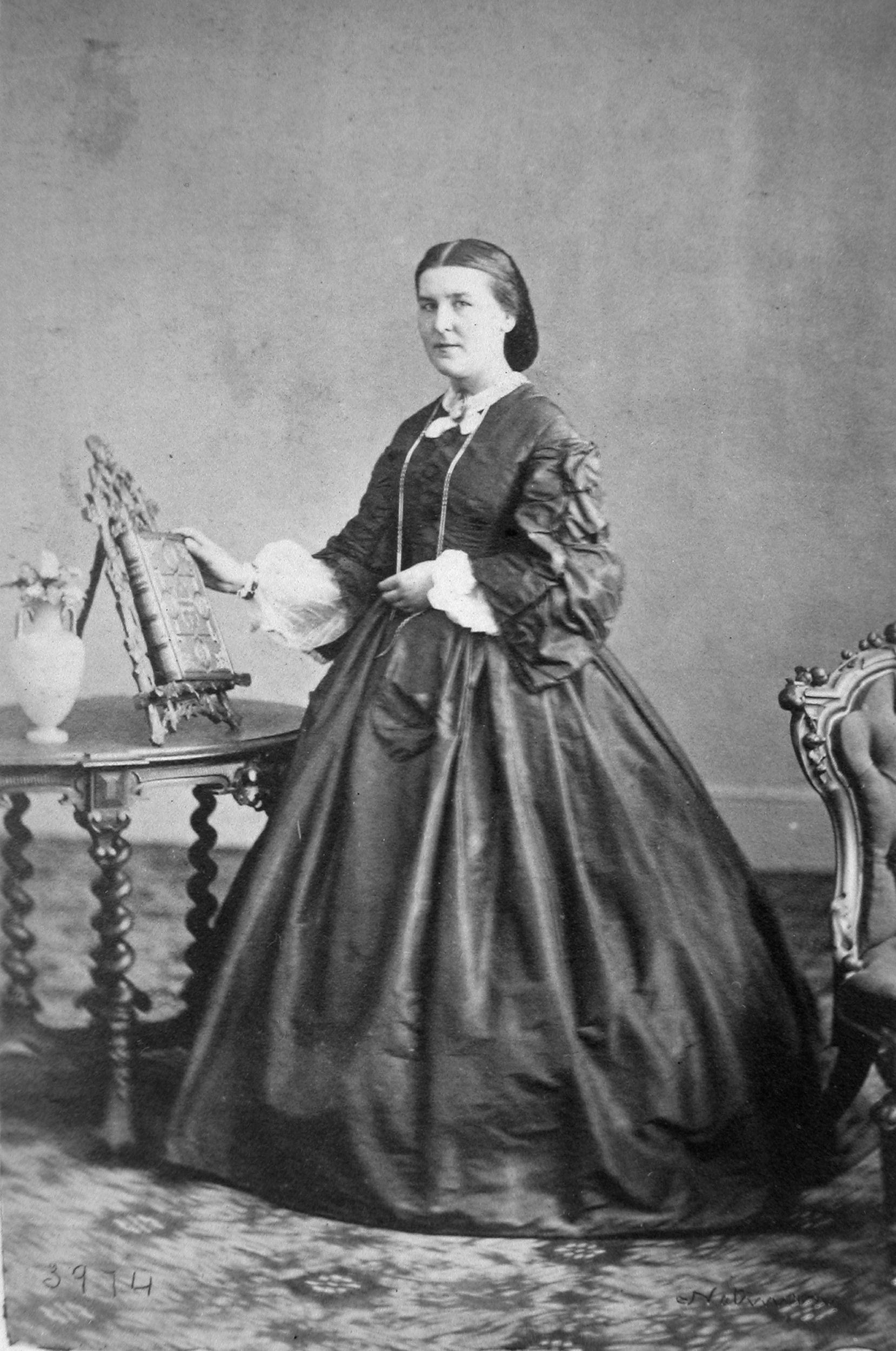 Black-and-white photograph of a white-appearing woman in a silk gown with a wide skirt, standing in front of a polished wood table with an elaborately bound book on a stand.