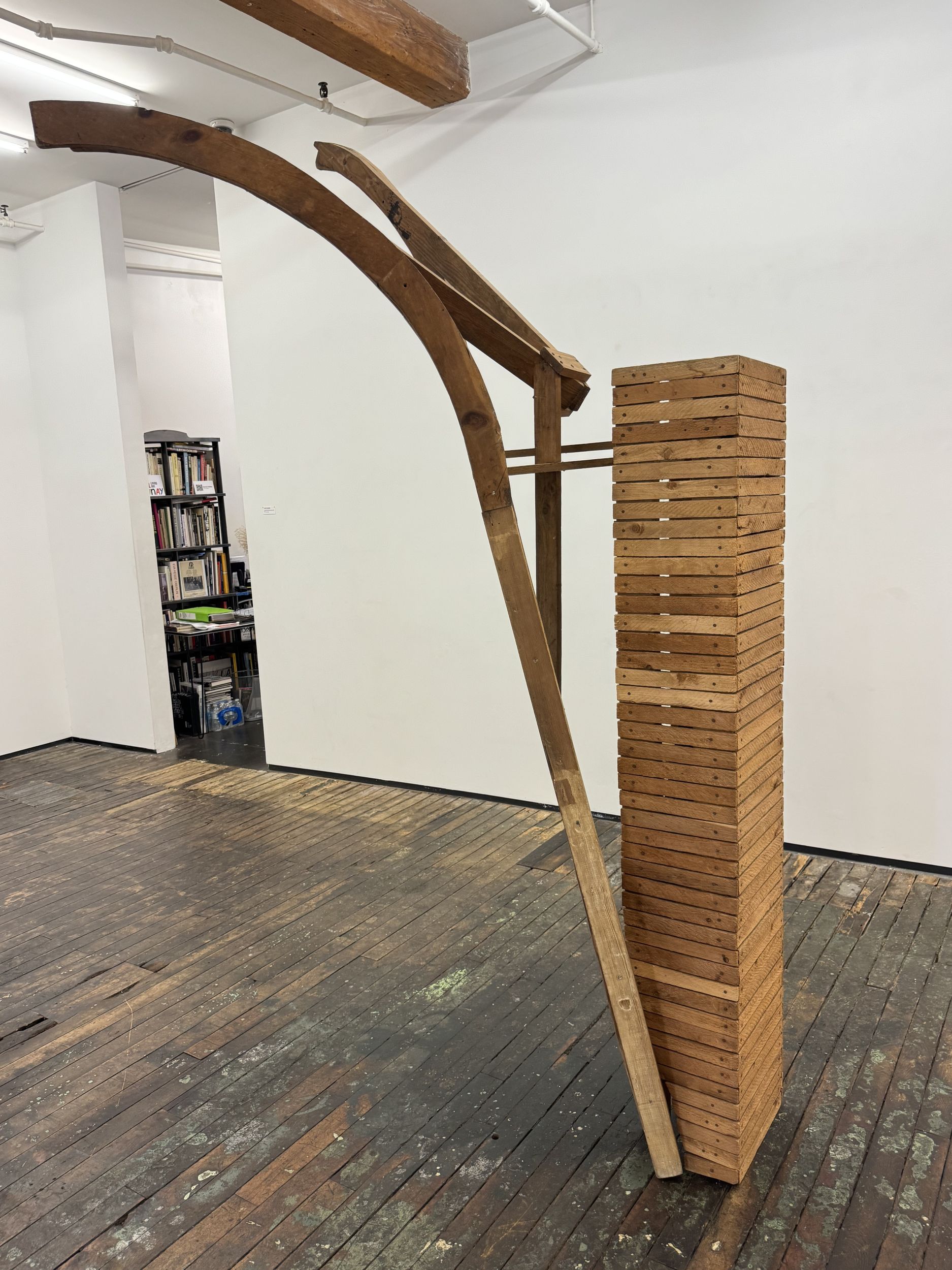 Wooden sculpture in a gallery setting. the sculpture is made of wooden slats and is rectilinear in form, with additional pieces that extend from its main form.