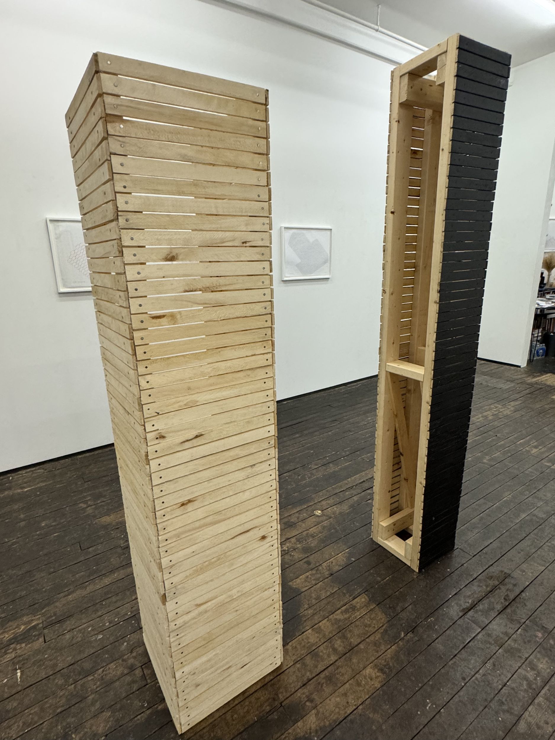 A pair of wooden sculptures in a gallery setting; the sculptures are vertical towers in form, made of wooden slats. The one on the right is partially painted black.