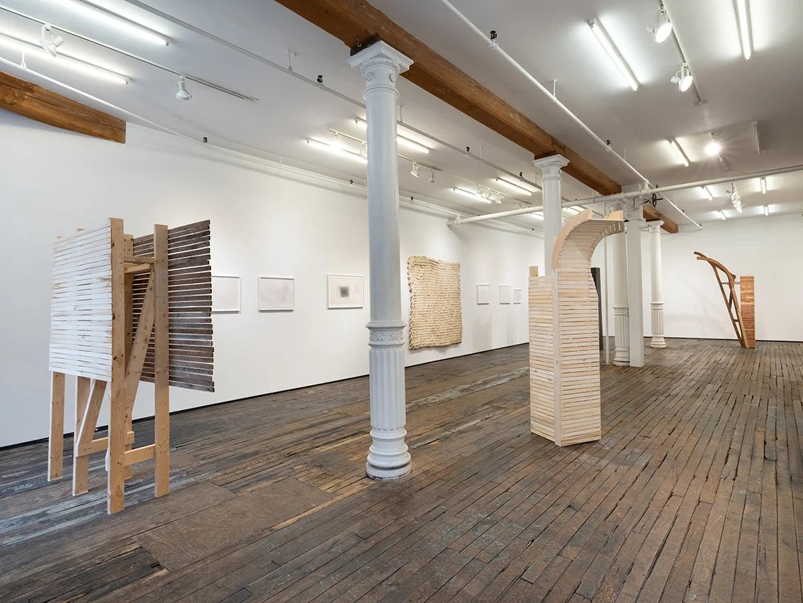 Interior of a museum gallery with rough wood floors, white painted walls, and classical columns supporting a central wooden beam. Wooden sculptures made of slats are scattered throughout the gallery.