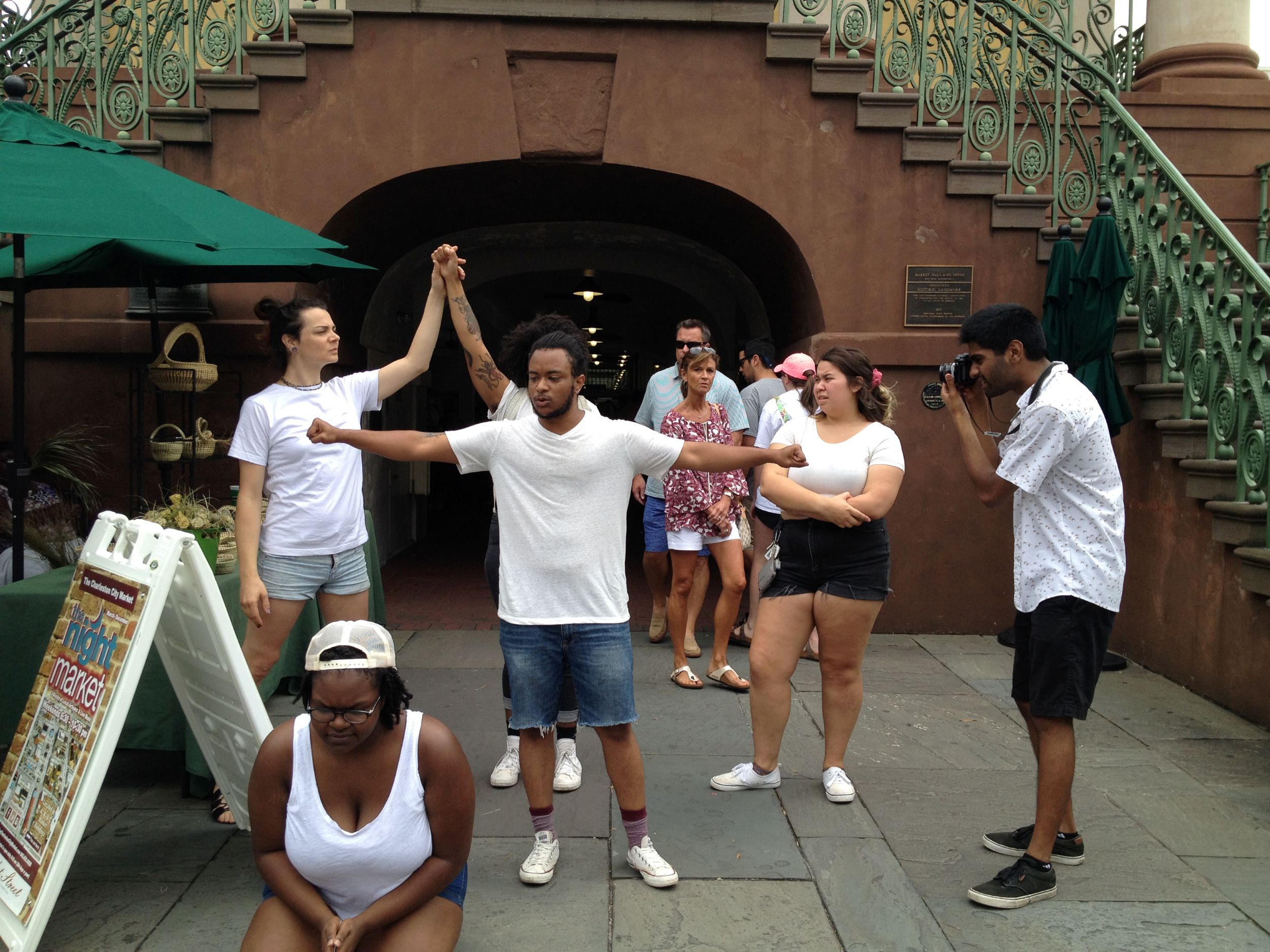 View of several people gathered in a courtyard conducting a performance. One woman crouches at the front, her hands clasped. A Black Man stands in the center, his arms outstretched and fists clenched. Two people in the background join hands over their heads while a white woman looks on with an expression of scorn. A man on the right takes a picture, while others observe.
