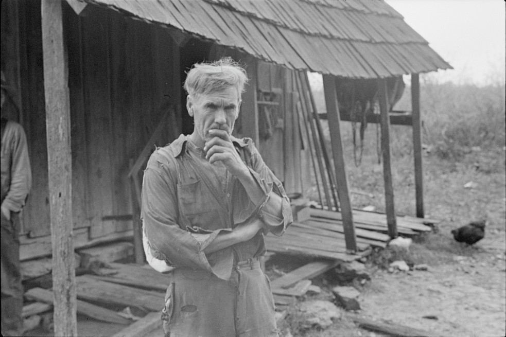 Black-and-white photograph of a white man in tattered coveralls standing in front of a wooden shack that is in disrepair. He holds his hand to his mouth in a gesture of concern.