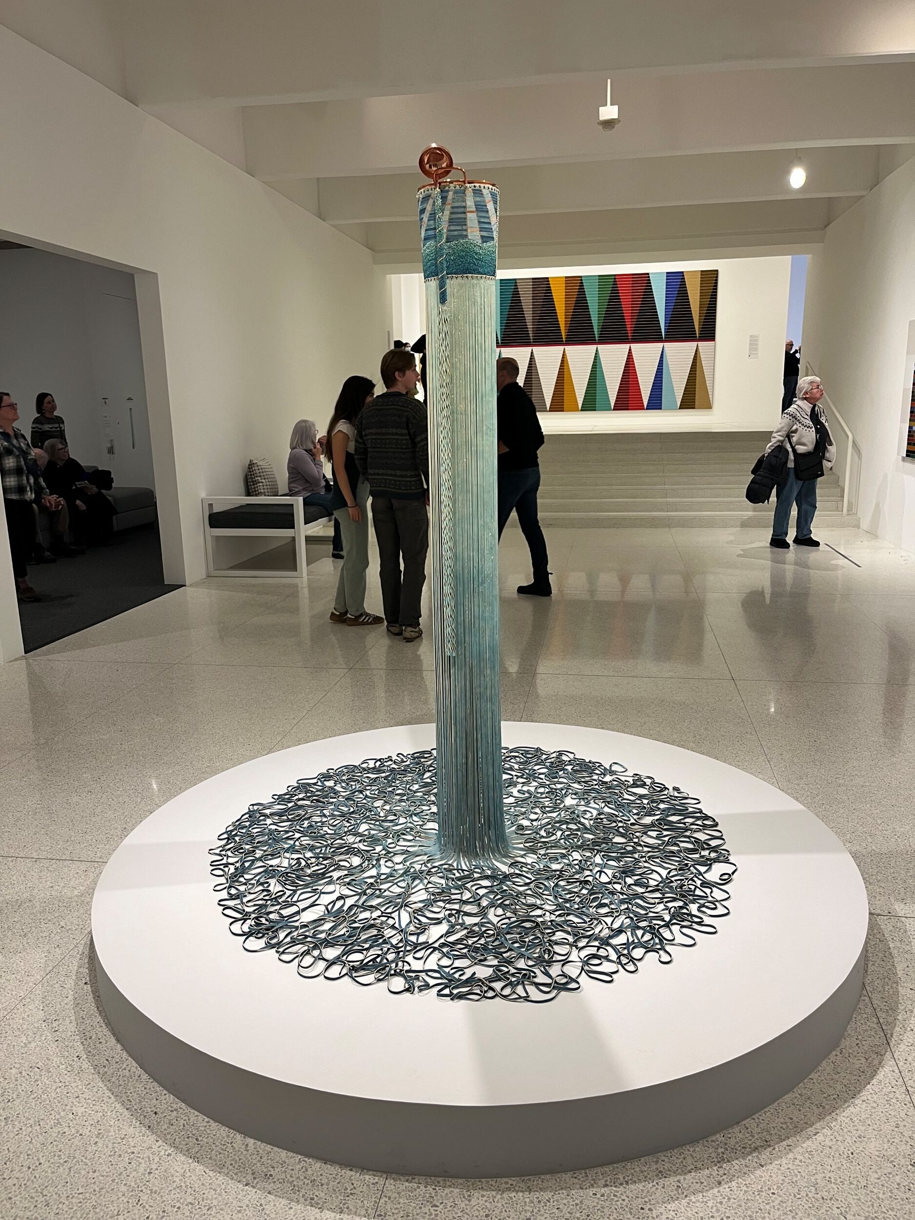 Interior of a museum gallery with a large sculpture on a round plinth in the foreground. The sculpture is in the form of a tall, pale green pole, and at its base it seems to dissolve into tangled threads.