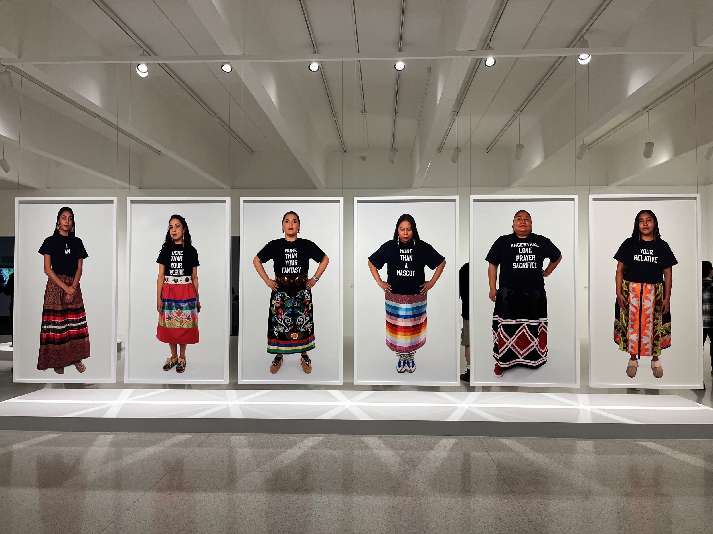 Interior of museum gallery showing six large framed images of women in long, colorful skirts, each wearing a black t-shirt with white lettering.