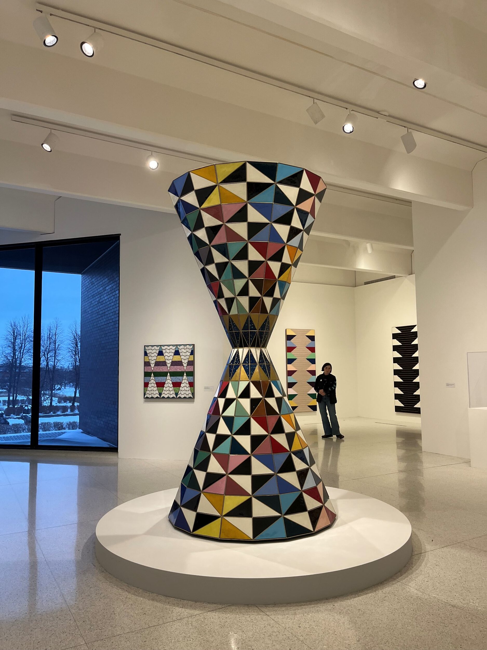 Interior of a museum gallery with a large sculpture standing on a round plinth in the foreground. The sculpture is hourglass-shaped and composed of multicolored triangles.