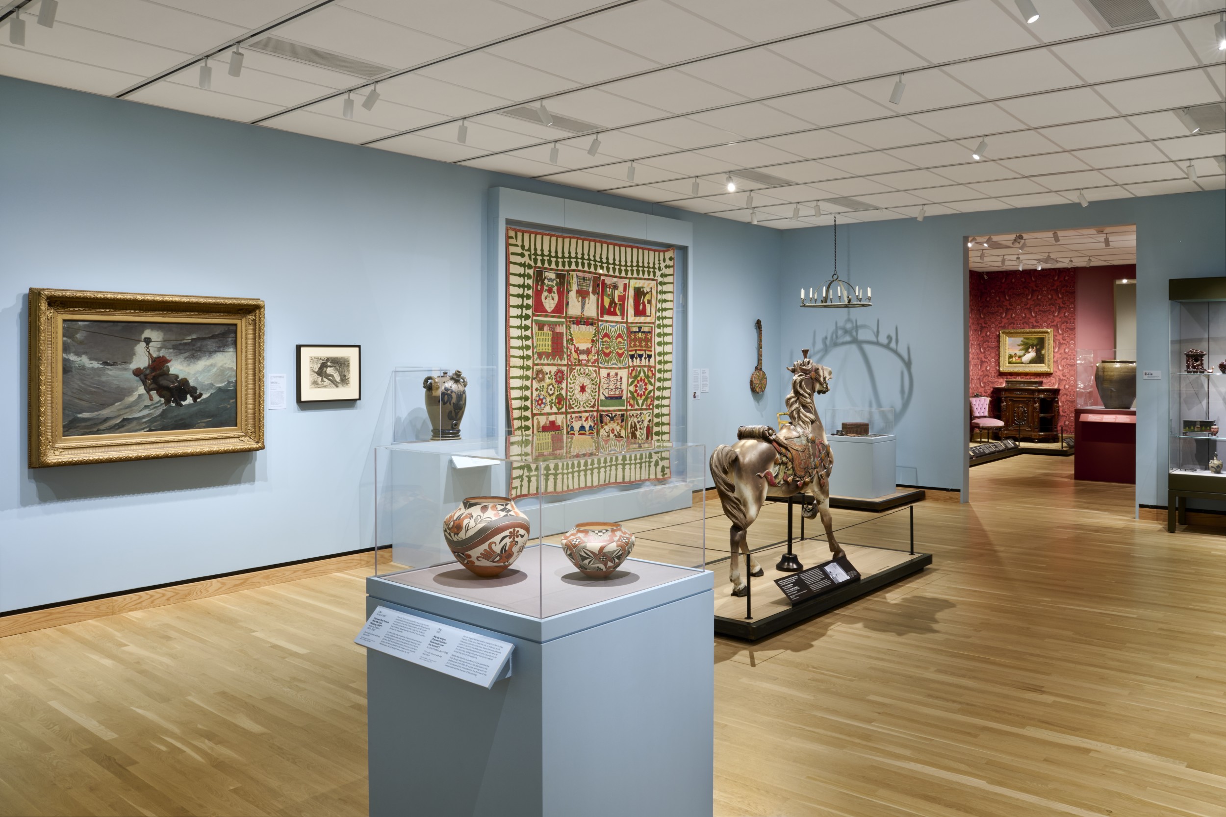 Interior of museum gallery with light blue painted walls. In the center of the room is a carousel horse; just in front of it are two Native American ceramic vessels in a case. On the wall to the left are two framed artworks, a sculpture in a case, a quilt, and what looks like a musical instrument.