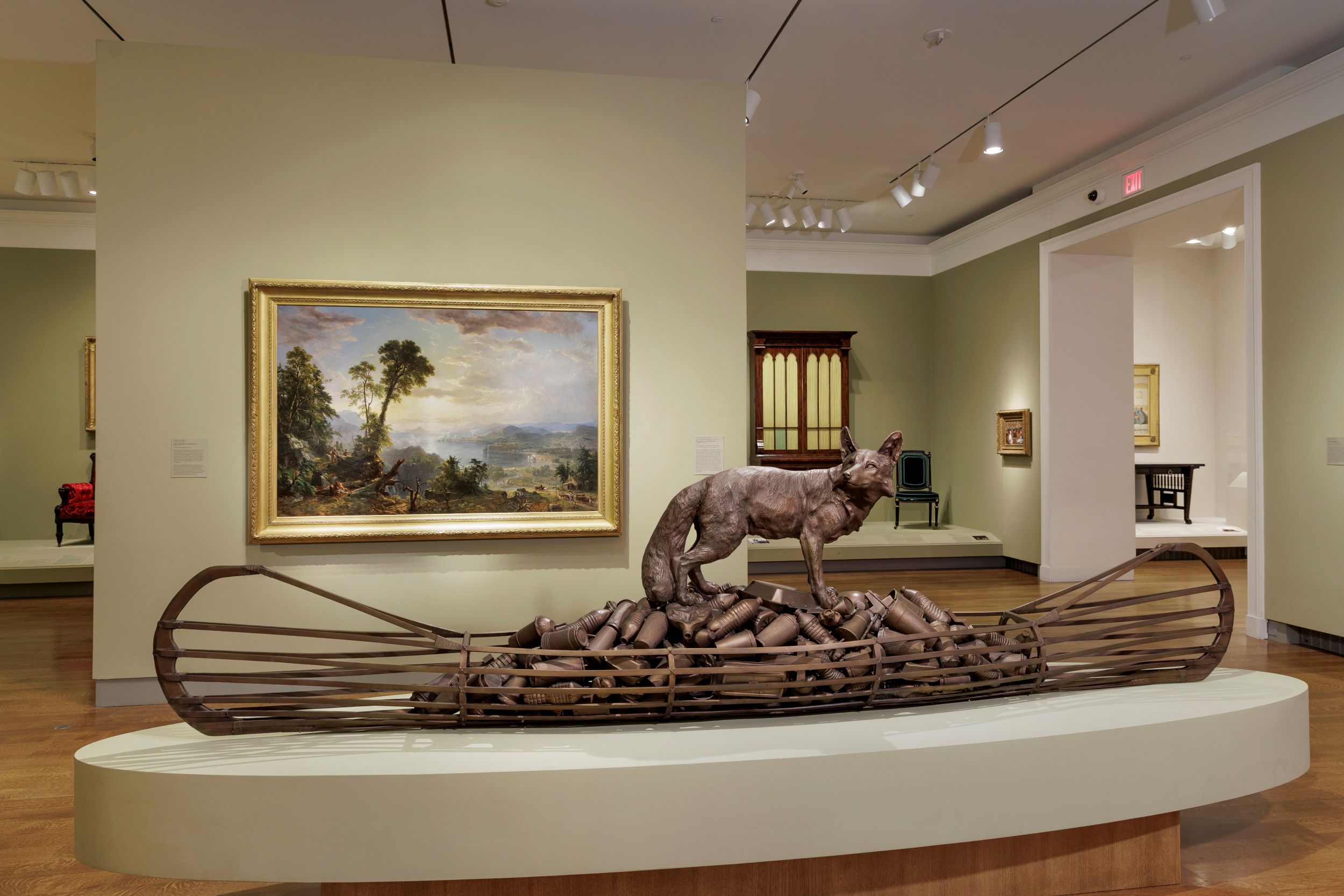 Interior of a museum gallery showing a bronze sculpture of a coyote standing on a pile of an unidentifiable material in the framework of a canoe. Behind him, on a freestanding wall, is hung a classically composed landscape painting.