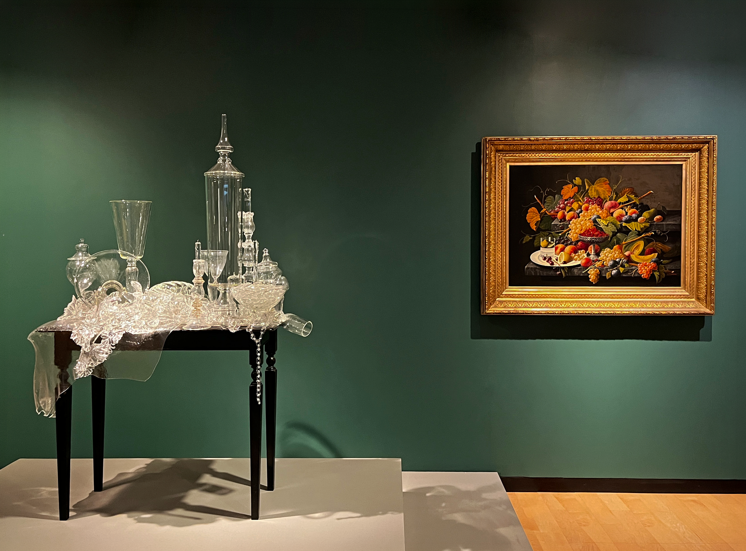 Head-on view of a museum gallery wall painted dark green, on which is hung a framed still life of fruits. To its left is a table covered with clear ornamental glass objects.