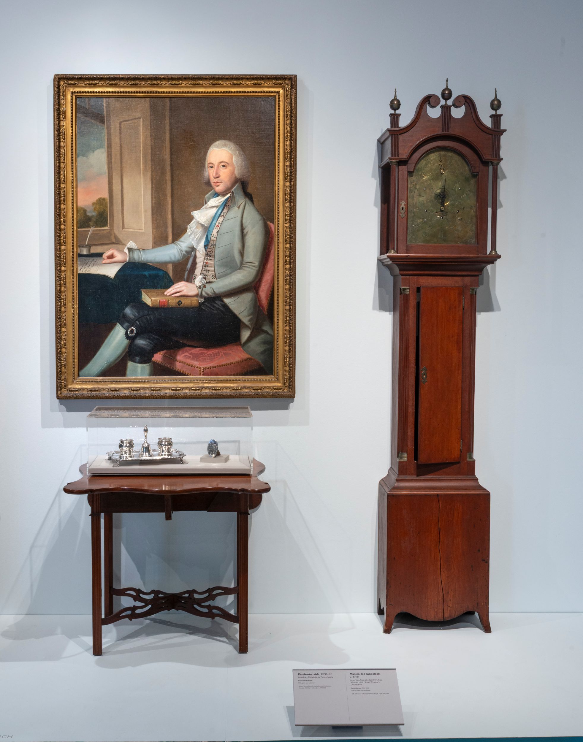 Head on view of a museum gallery, showing a tall-case clock on the right, and a table to the left, both resting on a plinth. Atop the table is a group of silver objects, and above it is a portrait of a man in black breeches and a gray waistcoat, sitting at a desk in front of a window.