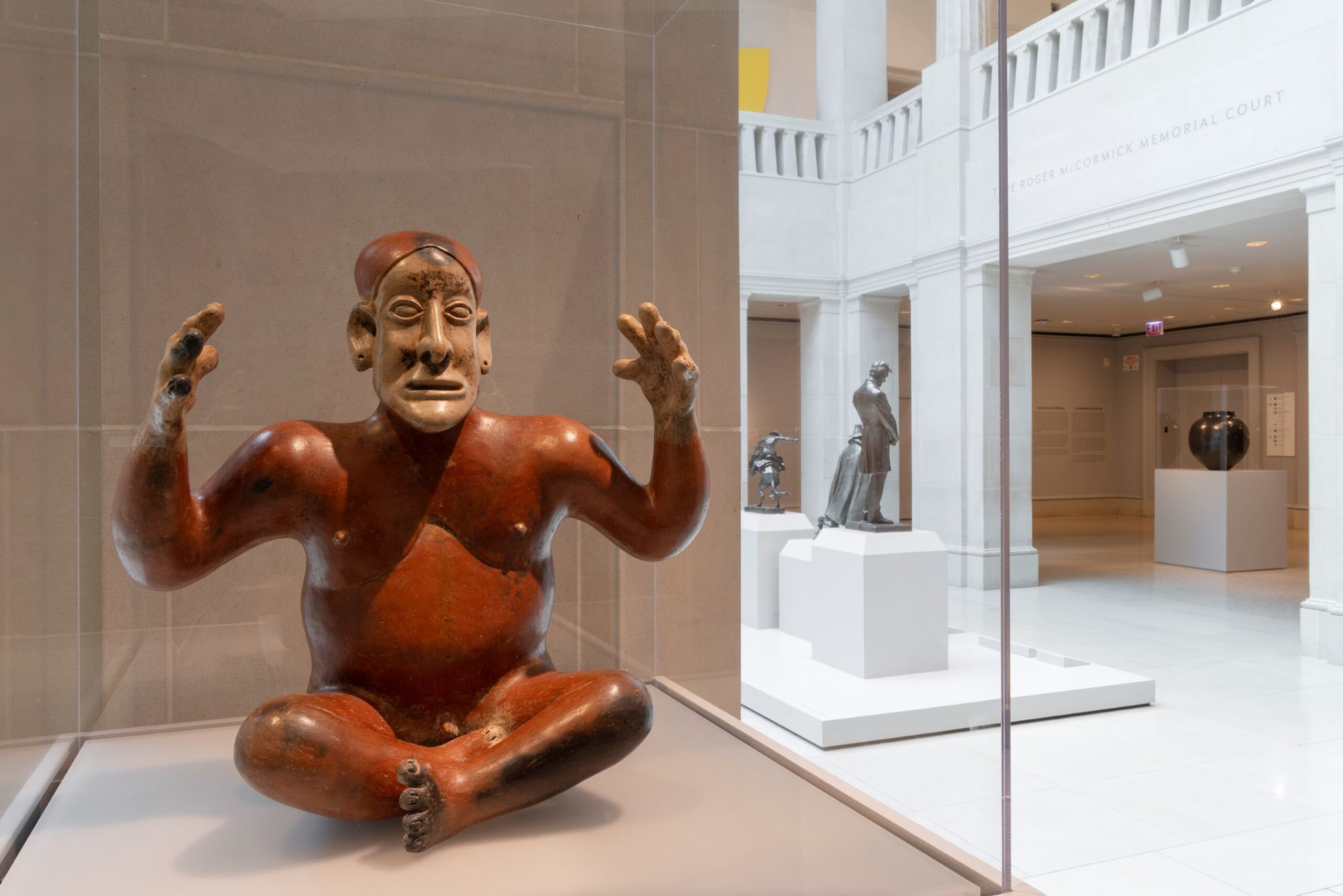 Meso-American pottery figure, sitting cross legged, with its hands raised above its shoulders, inside a museum case, with bronze sculpture and another pottery vessel visible in the background.