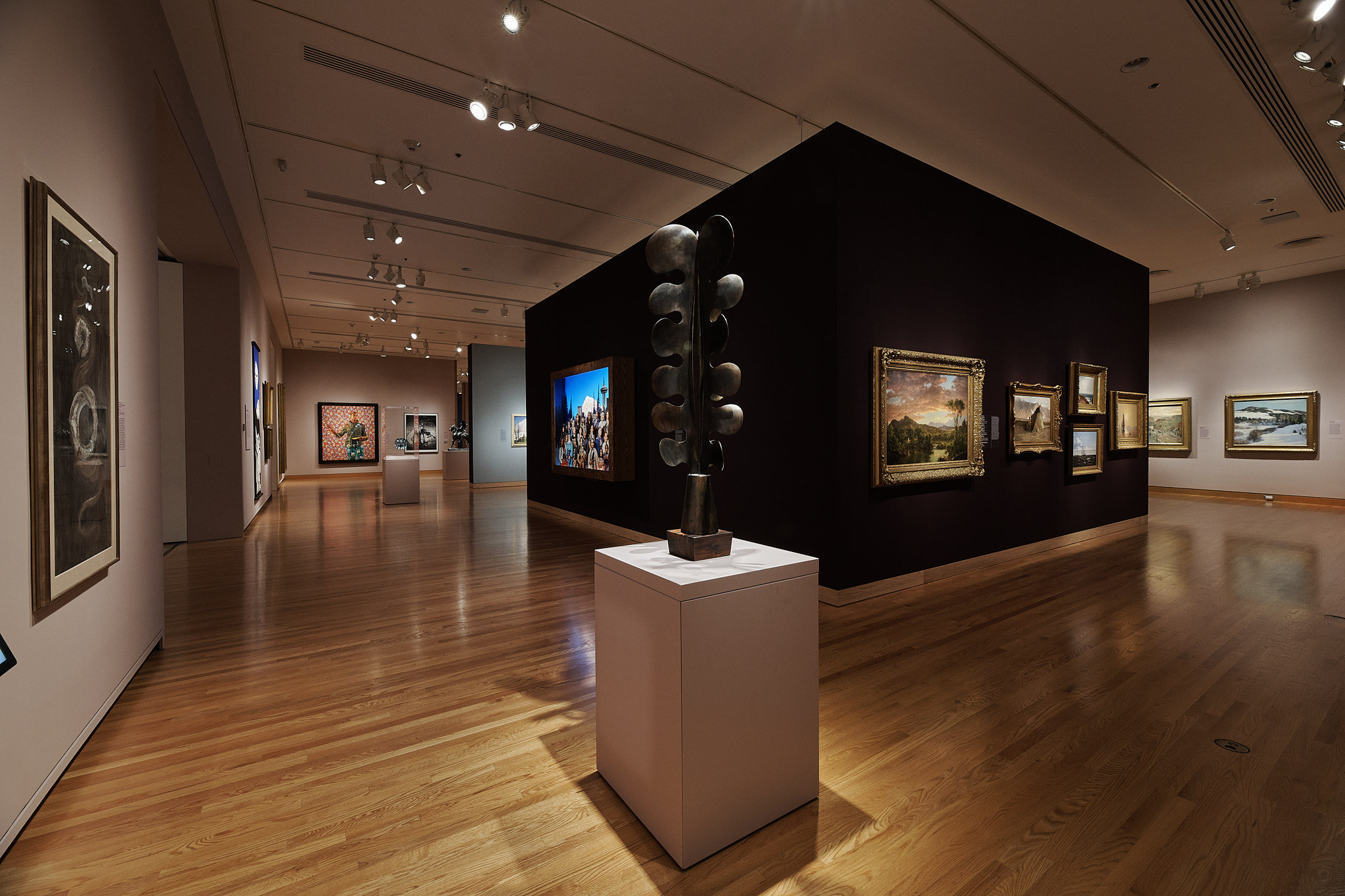 Interior of a museum gallery, showing a crossing of walls hung with framed works of art. Standing on a pedestal at the crossing is an abstract bronze sculpture in a cactus-like form.