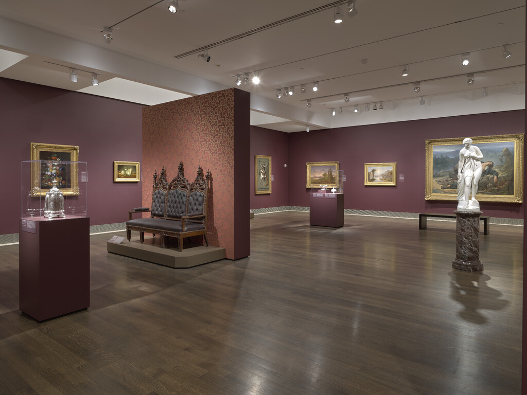 Interior of museum gallery with dark wood polished floors and dark red painted walls. Paintings hang on the walls, and on the floor are is a marble sculpture and two freestanding cases of decorative arts. An ornate loveseat sits against a freestanding half-wall.